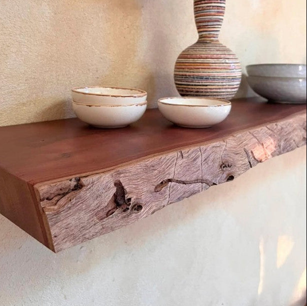 A rustic floating wooden shelf mounted on a textured beige plaster wall, styled with a striped ceramic vase holding pink flowers, two small white bowls, and a larger gray bowl.
